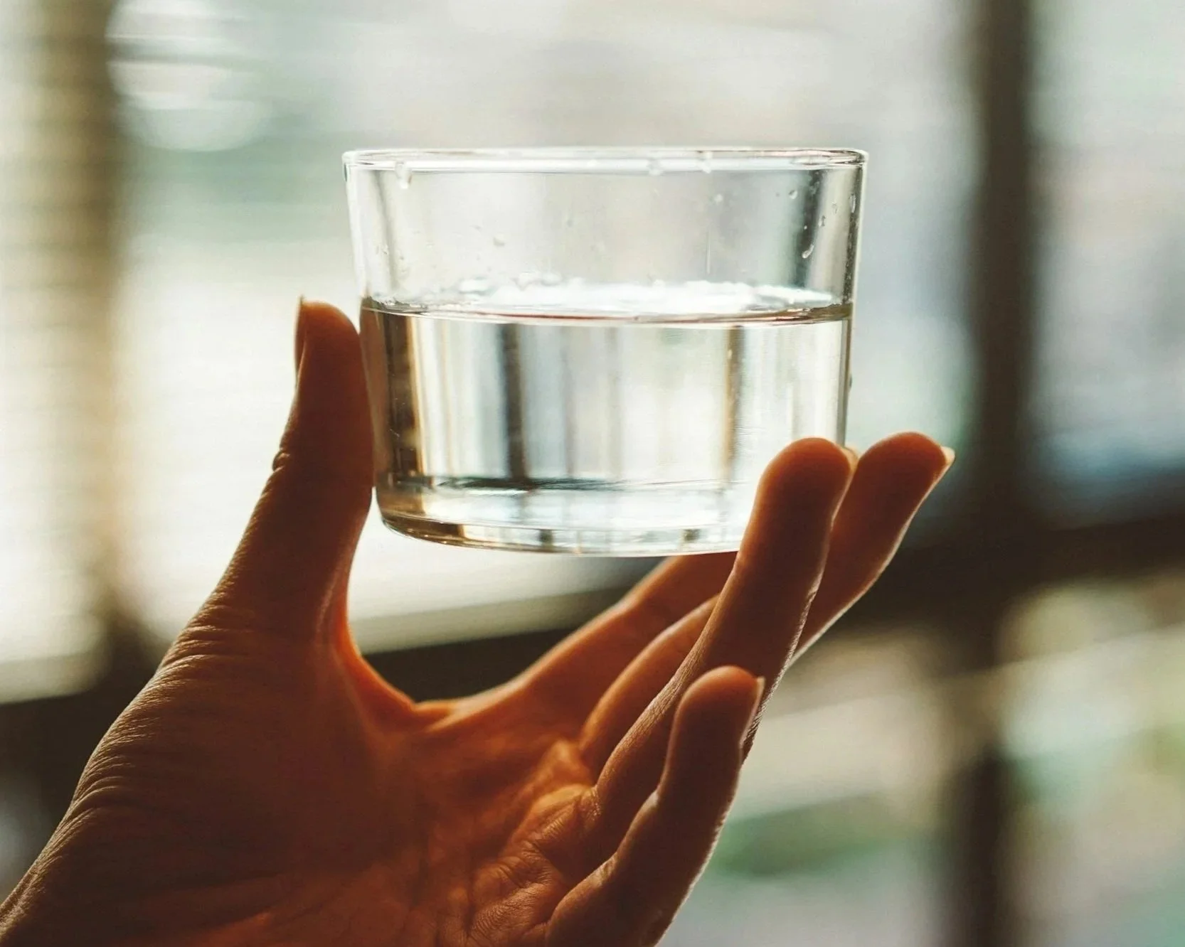 A hand holding a glass of water against a blurry window background.