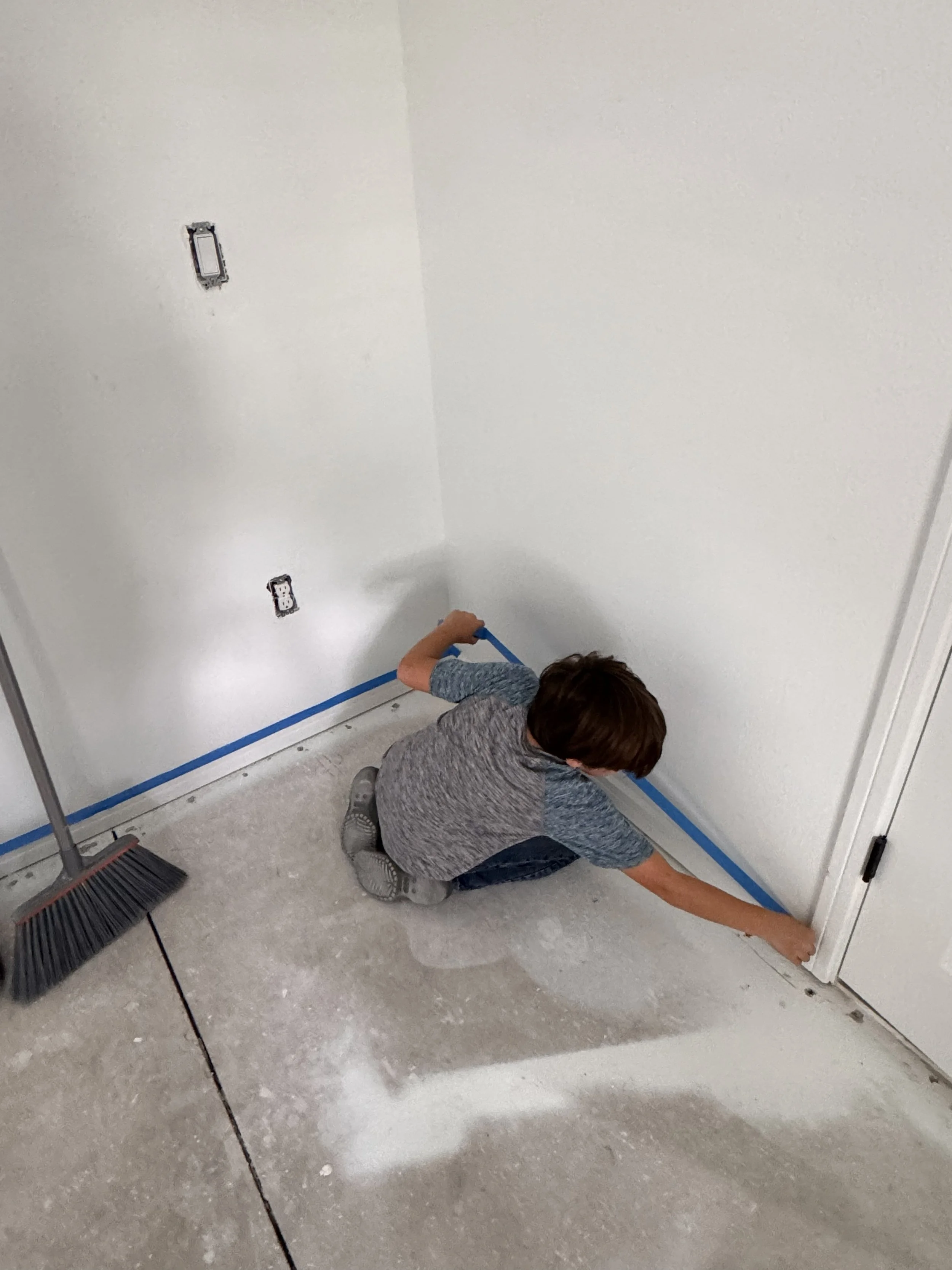 A young boy crouches on the floor, using painter's tape to create a border along the bottom of a white wall during home renovation.
