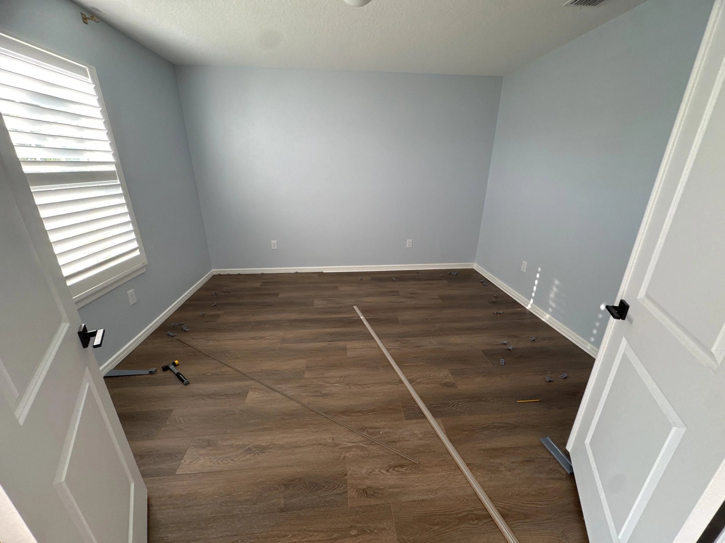 Empty room with light gray walls, wood flooring, and a window with white blinds. Construction tools and materials are on the floor, indicating ongoing renovation.