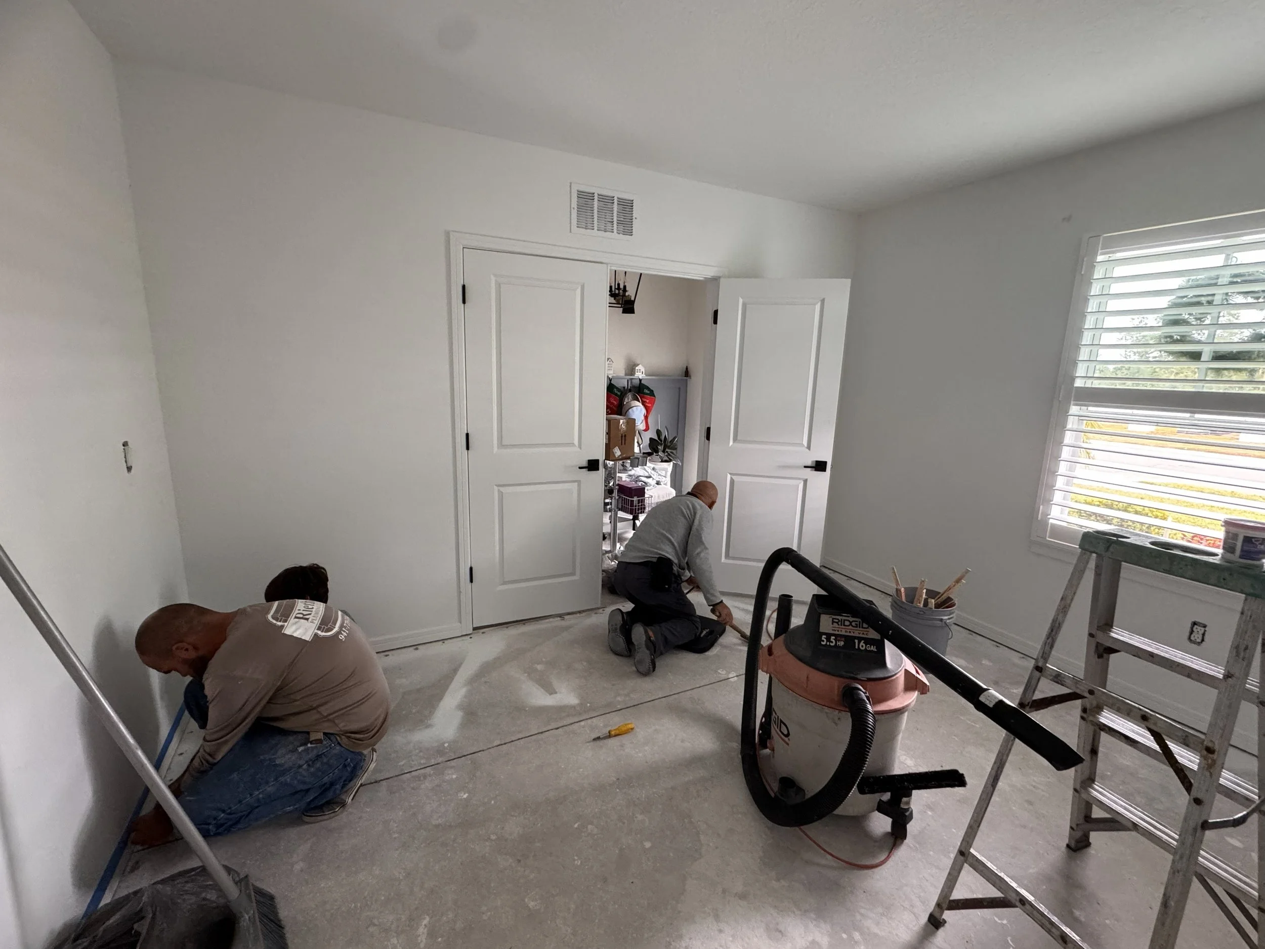 Two workers installing baseboards in a room during home renovation, with a ladder, a vacuum, and construction tools visible.