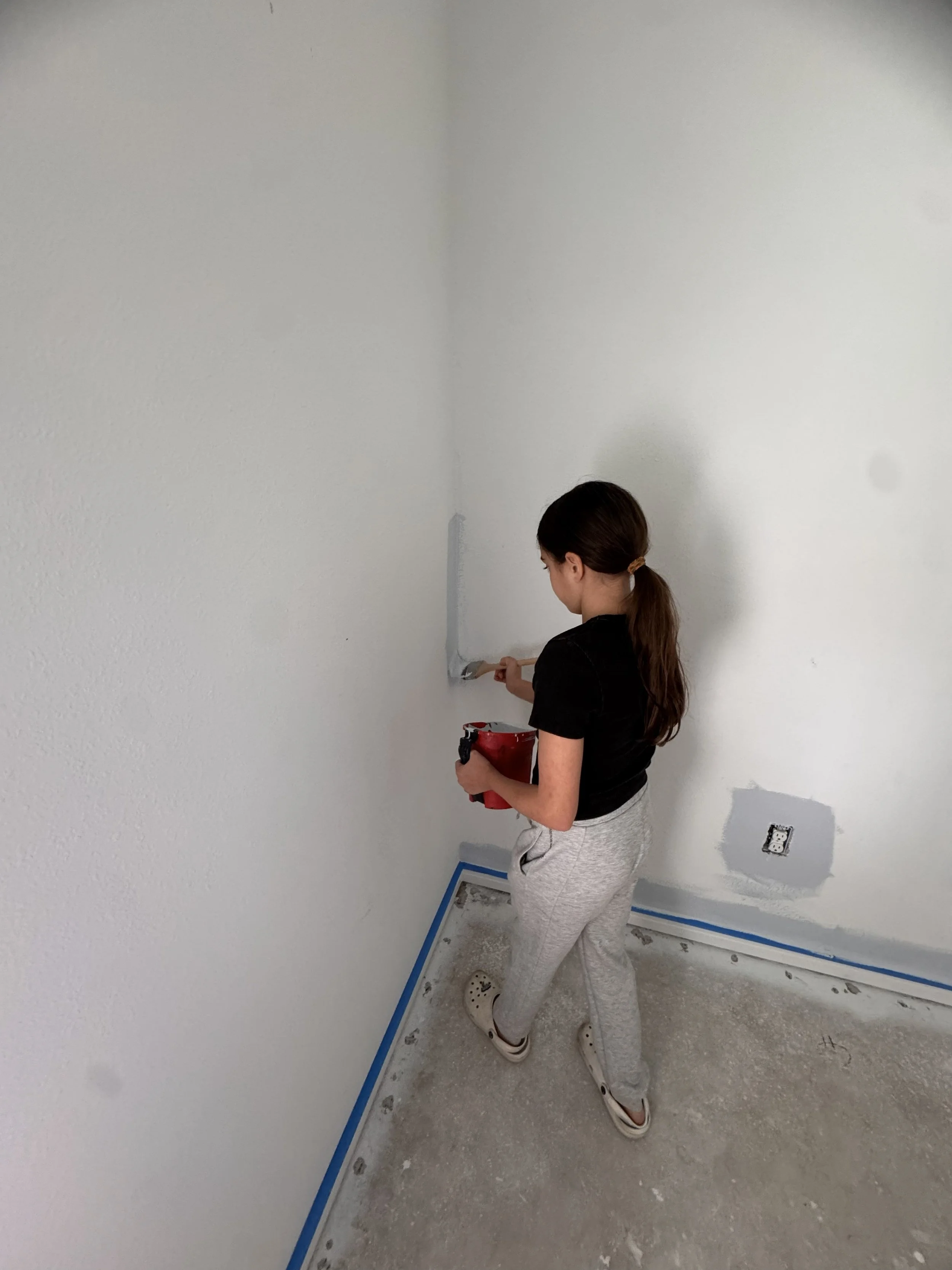 A girl painting a corner of a white wall in a room, holding a paintbrush and a red paint container, with blue painter's tape along the edges of the floor.