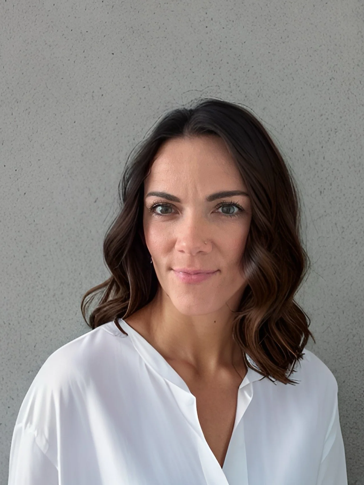 A woman with wavy brown hair and light eyes wearing a white top standing against a plain, light-colored wall.