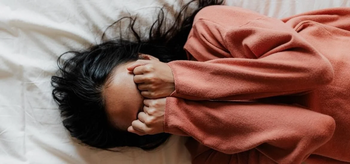 A woman lying on a bed, covering her face with her hands, wearing a long-sleeved, rust-colored top.