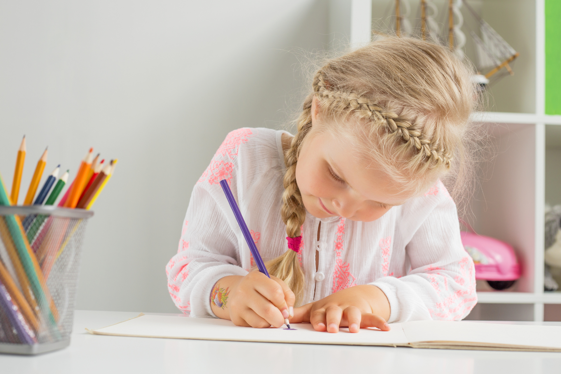 A young girl with braided blonde hair writing in a notebook at a white desk, with a container of colorful pencils on her left.