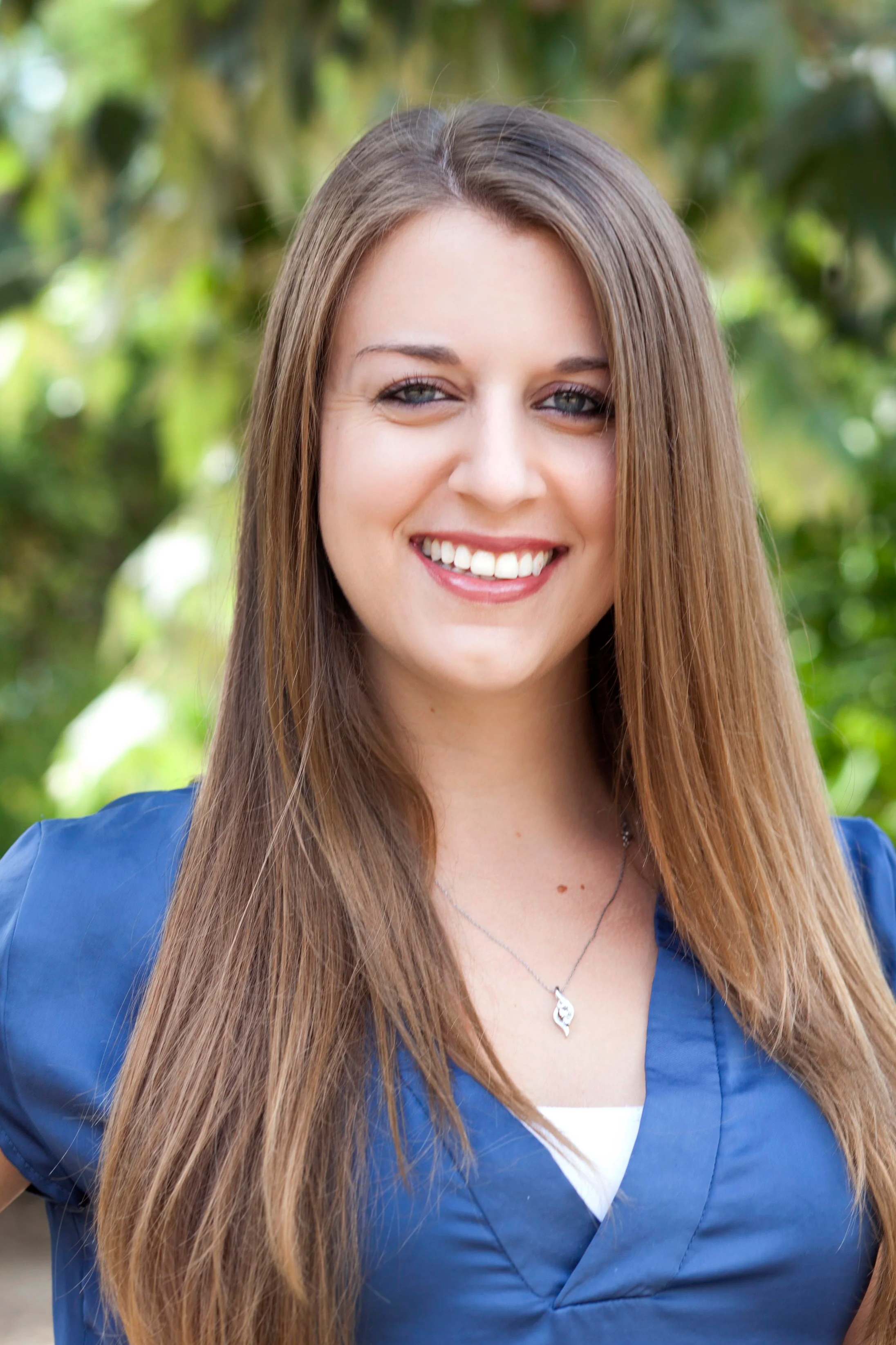 A young woman with long brown hair smiles outdoors, wearing a blue top and a silver necklace, with greenery in the background.