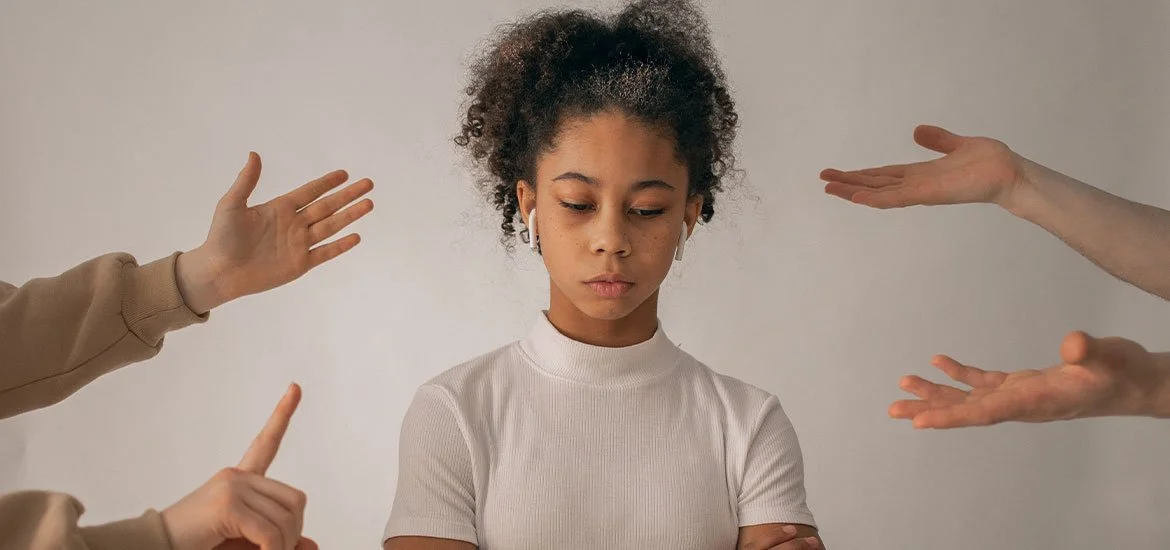 A young woman with curly hair, wearing a white shirt and wireless earbuds, looks down with a serious expression while multiple hands around her, some with different skin tones, gesture with open palms and pointed fingers towards her.