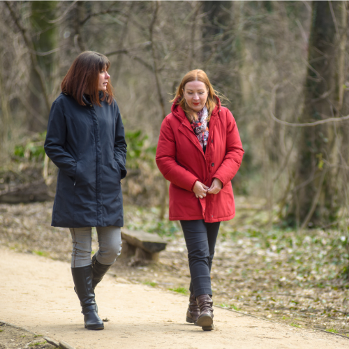Two women walking on a dirt path in a wooded area, engaged in conversation, one wearing a navy coat, the other a red coat.