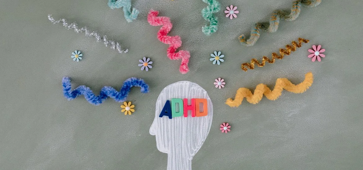 Decorative art featuring a paper cutout of a human head with colorful foam letters spelling 'ADHD' on the forehead, surrounded by colorful pipe cleaners and small flower stickers on a light gray background.
