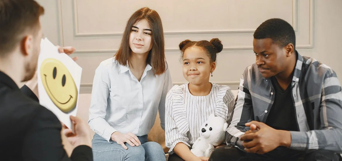 Group therapy session with a person holding a paper with a smiley face, three young people sitting and listening, including a girl holding a stuffed polar bear.