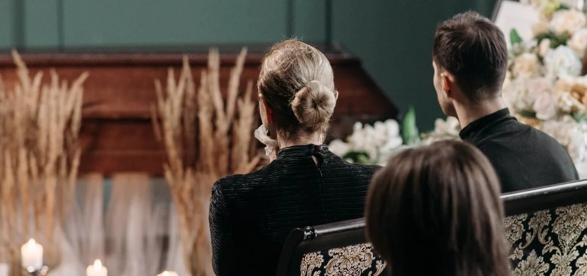 People sitting on a vintage floral couch, facing an altar with flowers and candles, during a ceremony.