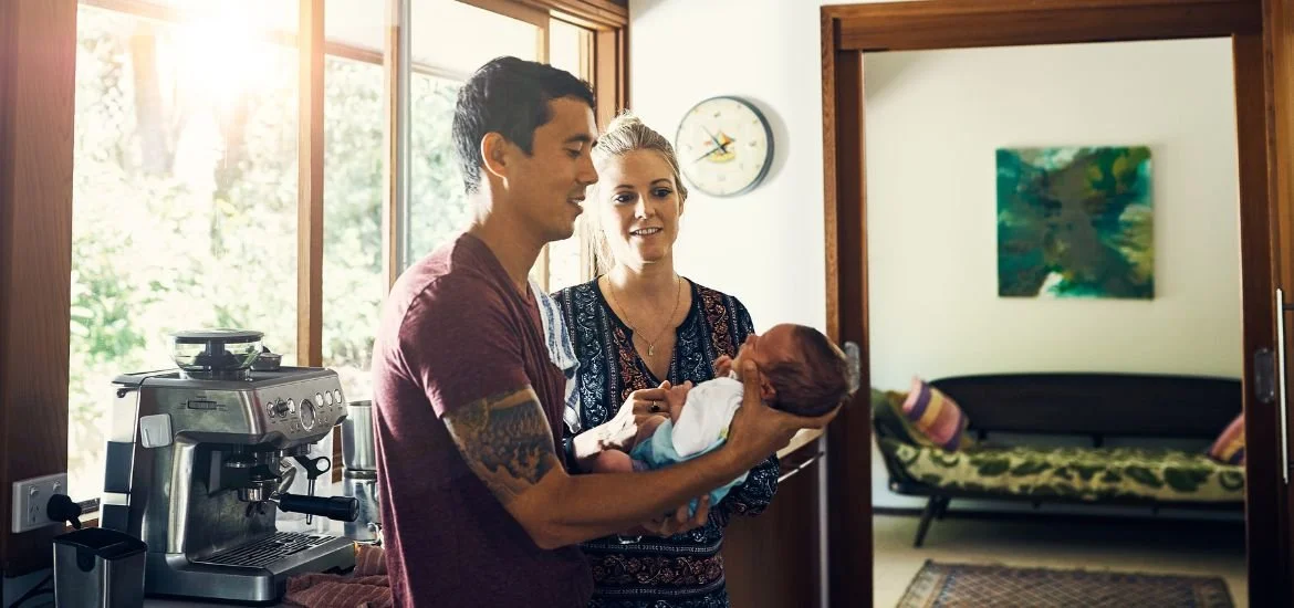 A couple holding a newborn baby inside a home, with sunlight coming through a window and a coffee machine nearby.
