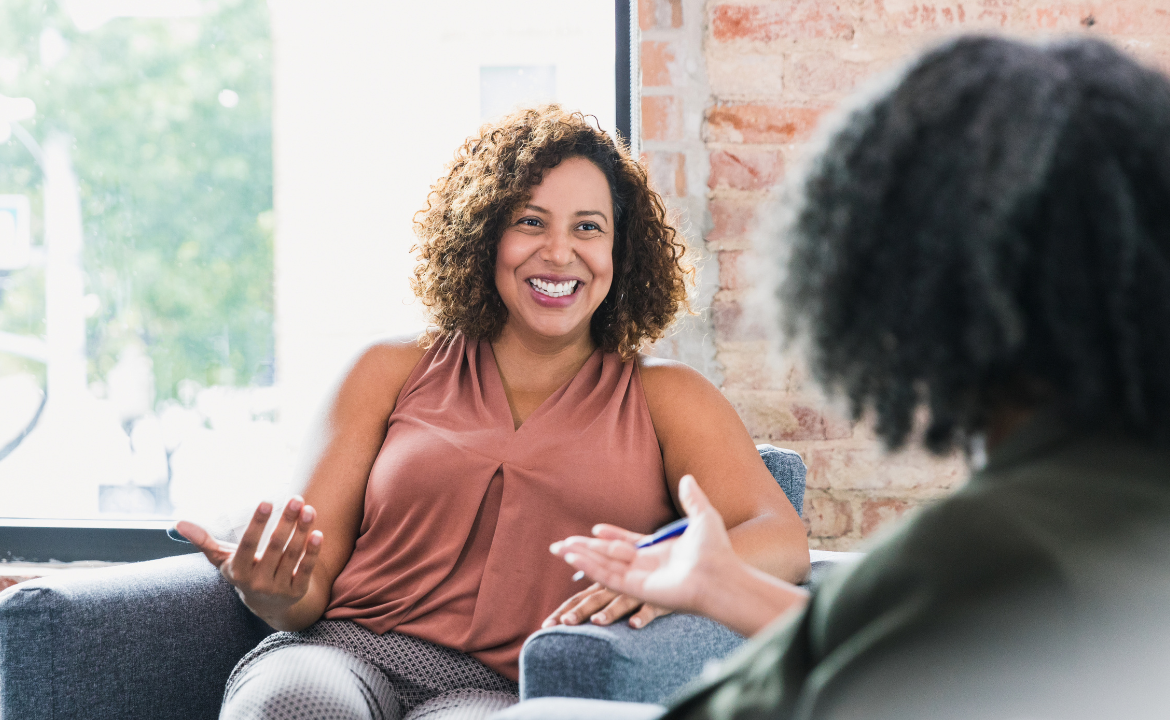 Two women having a conversation in an indoor setting, with one woman smiling and gesturing with her hand.