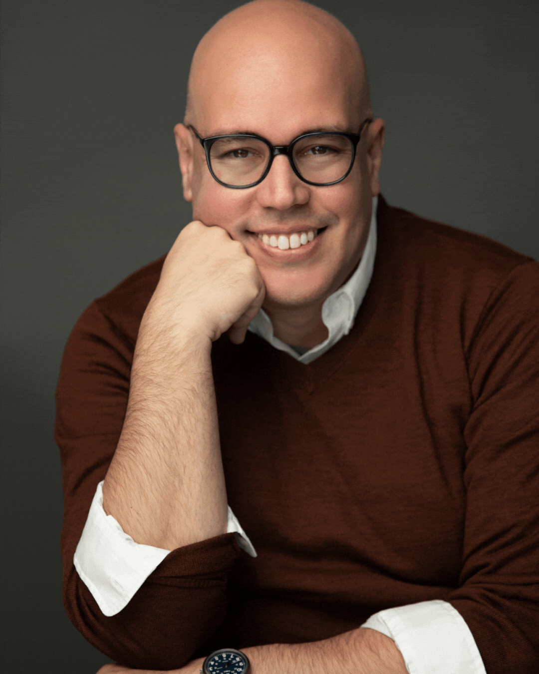 Portrait of a smiling man with glasses, wearing a white shirt and brown sweater, resting his chin on his hand against a dark background. Robert W. Schneider.