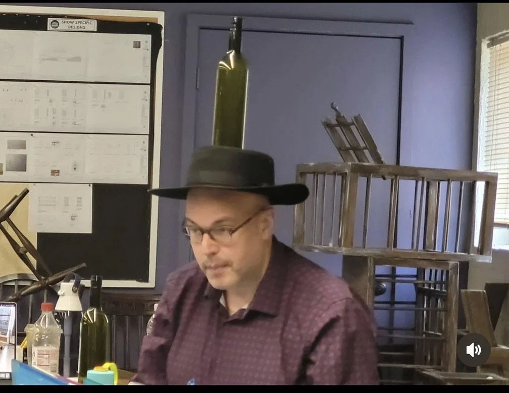 A man wearing glasses, a black hat, and a burgundy shirt sitting at a table with bottles and a laptop in a room with a display board and stacked folding chairs.