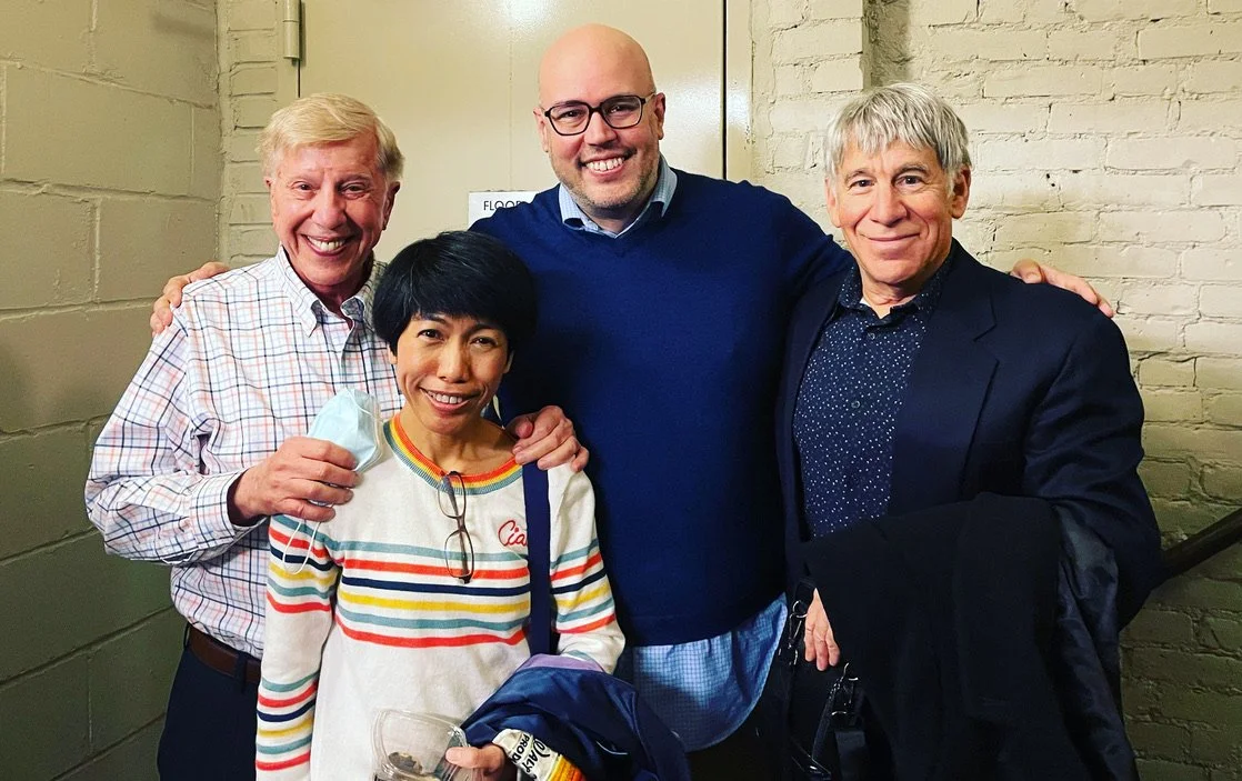 Four people smiling and posing together indoors, with one person holding a face mask and a drink, against a brick wall.