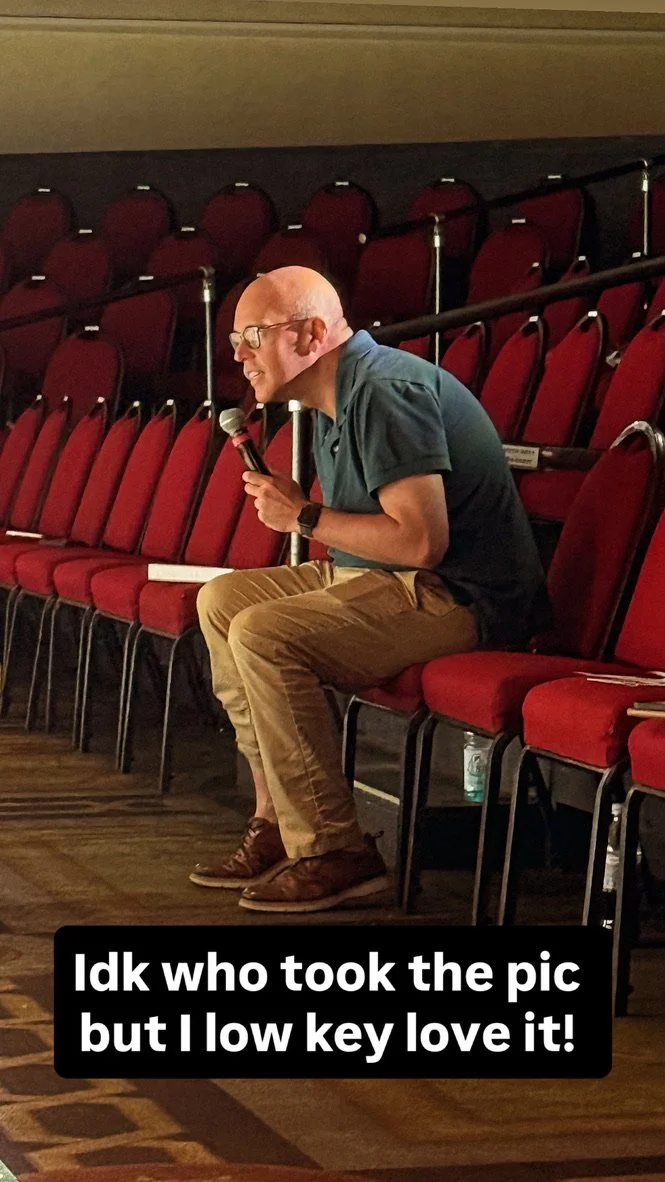 A man sitting alone in an auditorium with red chairs, holding a microphone and looking towards the stage, with a caption that reads 'Idk who took the pic but I low key love it!'