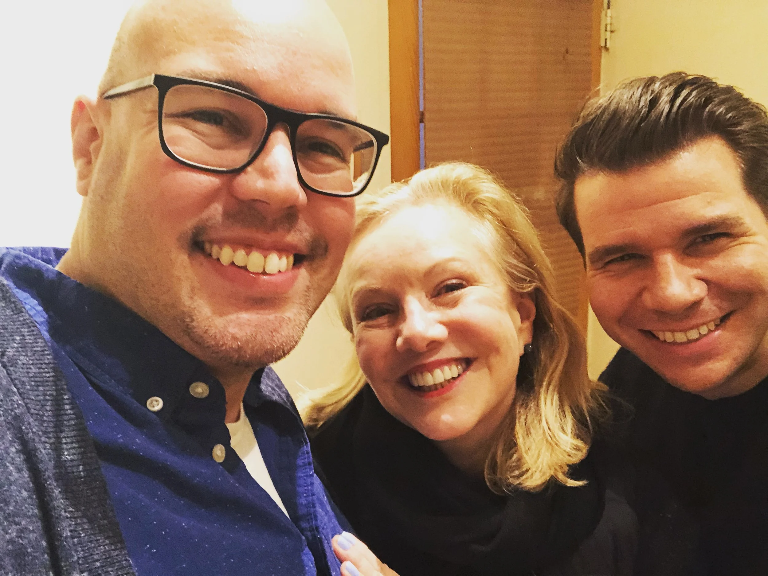 Three smiling people taking a close-up selfie, showing two men and one woman, indoors with a wooden background.
