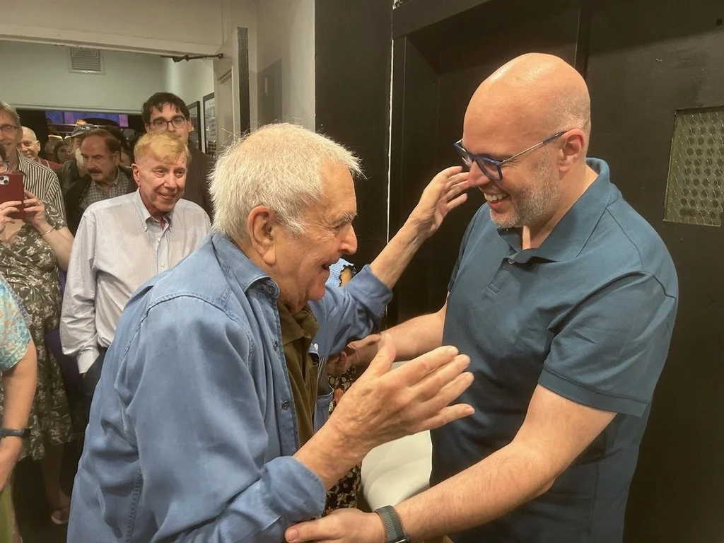 Two men, one elderly with white hair wearing a blue denim shirt, and one middle-aged with a shaved head and glasses wearing a dark blue polo shirt, smiling and engaging in a conversation, with several people observing in the background.