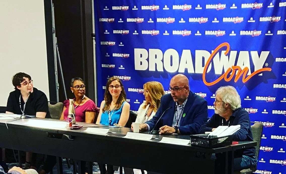 Six individuals seated at a panel in front of a blue backdrop with the word "Broadway Con" repeated on it. The panel includes a diverse group of men and women, some with papers and microphones.