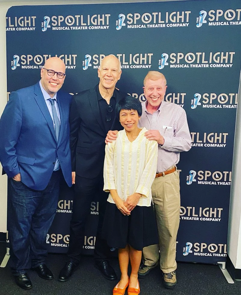 Four people smiling and posing together in front of a backdrop with 'The 2 Spotlight Musical Theater Company' logo, at a theater event.