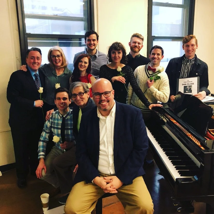 A group of eleven people, some holding white roses, posing in a room with large windows, with a grand piano in the foreground.