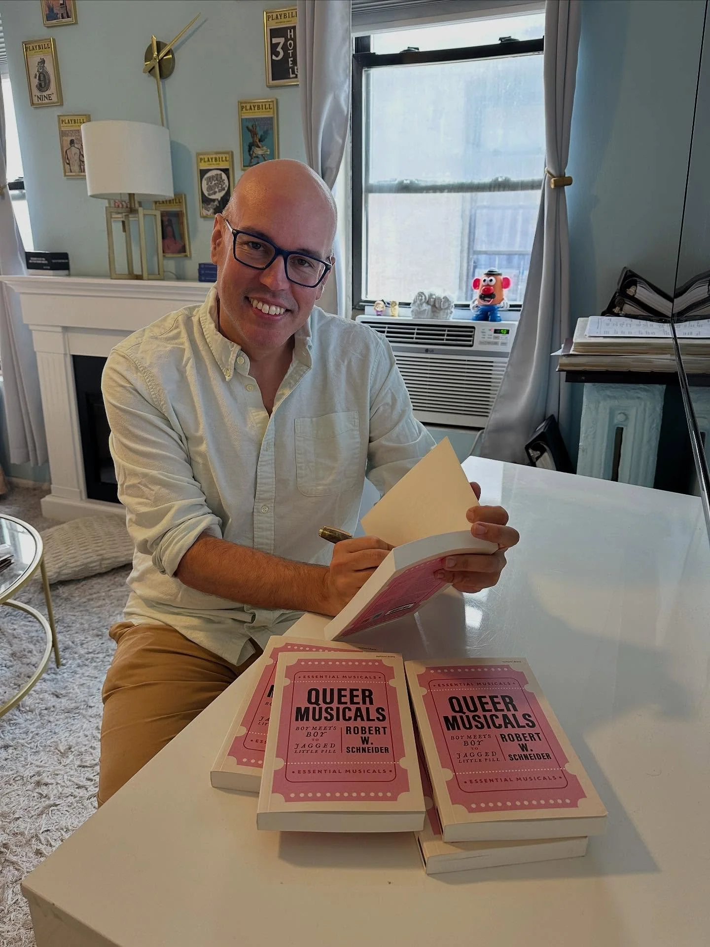 A man sitting at a white table, signing a book titled 'Queer Musicals' by Robert W. Schneider. Several copies of the book are stacked on the table. The man is wearing glasses and a light-colored button-up shirt. Behind him, a living room features a f
