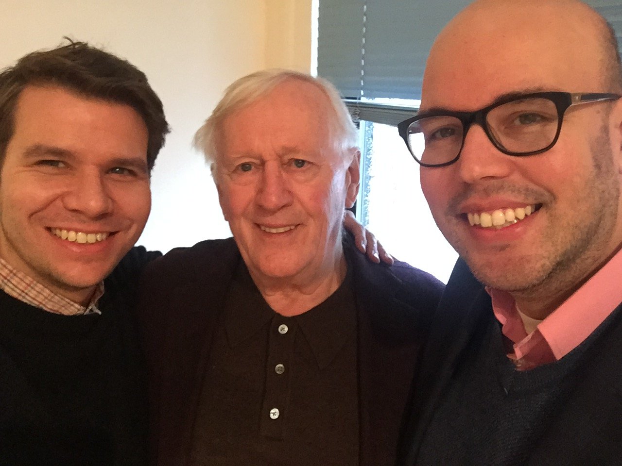 Three men smiling for a selfie indoors, with a window and blinds in the background.