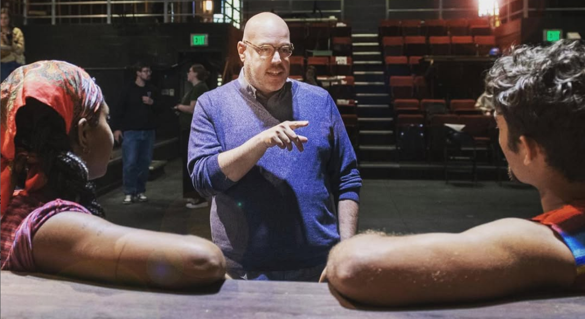 A bald man in glasses talking to two young athletes with arms resting on a table in a theatre or auditorium setting. Robert W. Schneider.