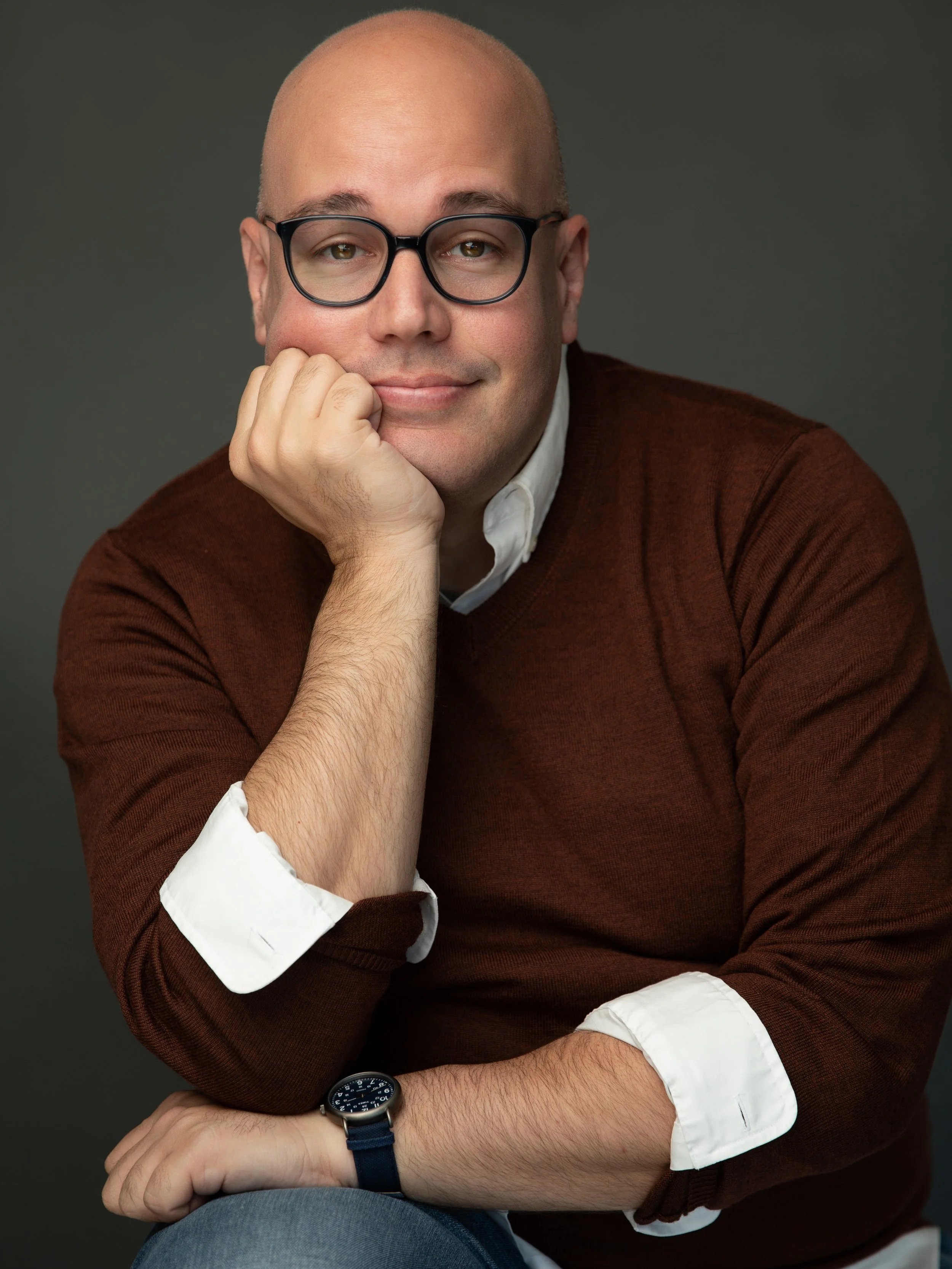 A man wearing glasses, a white collared shirt, and a brown sweater, sitting with his chin resting on his hand, smiling slightly at the camera against a dark background. Robert W. Schneider.