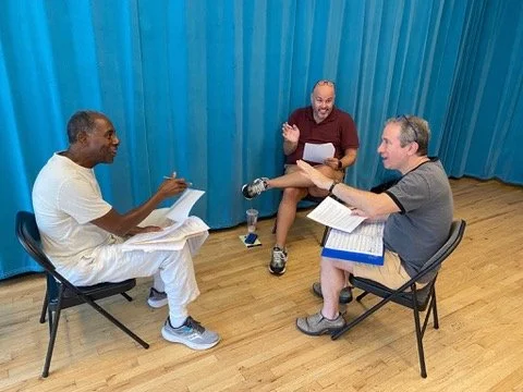 Three men sitting in a circle during a rehearsal or script reading, with scripts and notes, in a room with wooden floors and blue curtains.