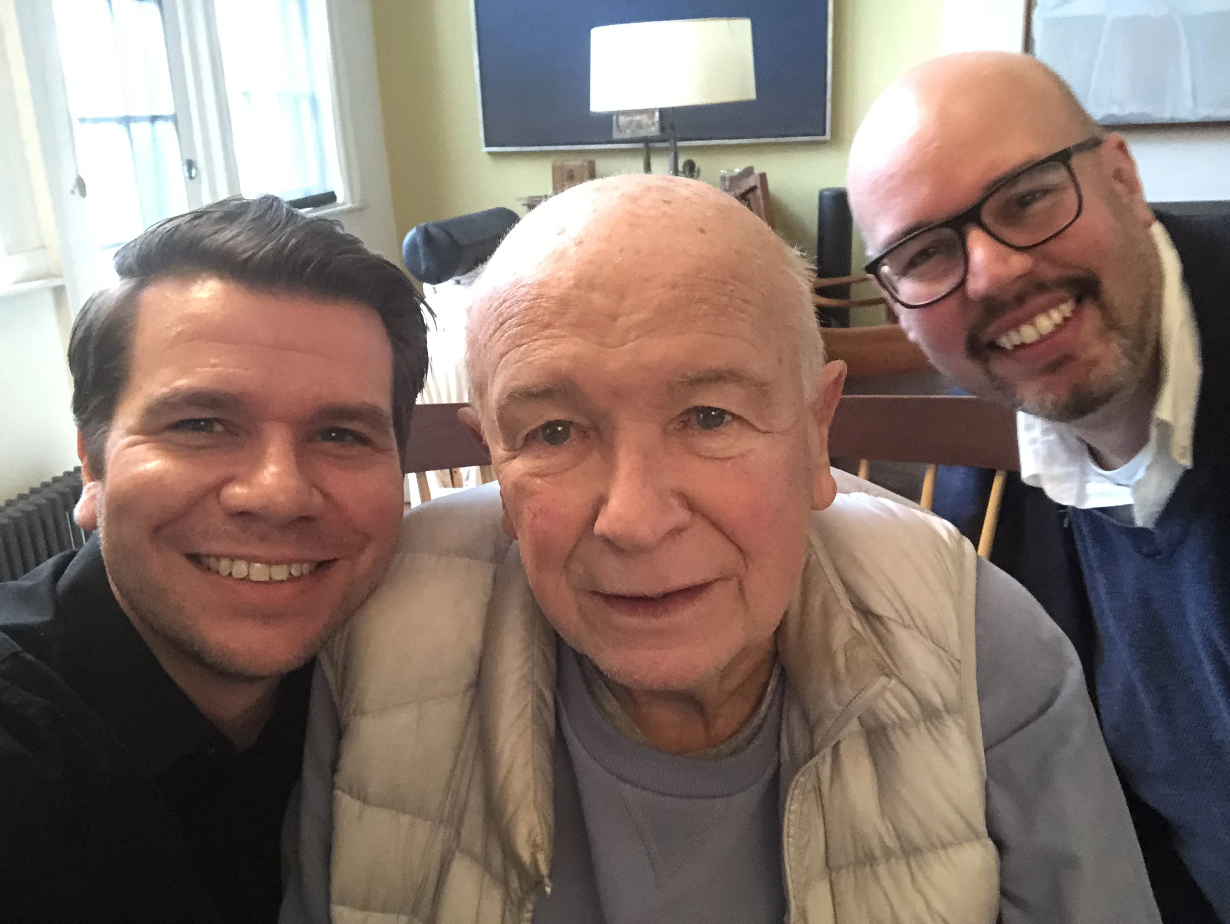 Three men smiling for a selfie in a room with a TV, a lamp, and windows in the background.