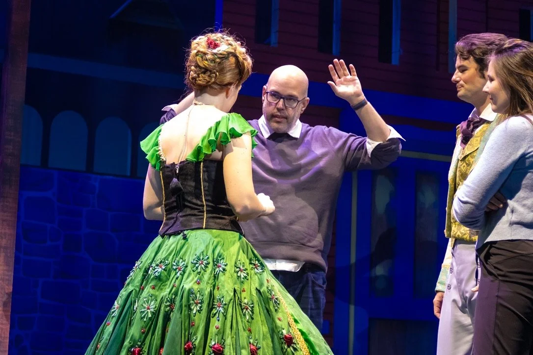 The image shows a theatrical scene with four actors. One woman dressed in a bright green, ornate dress with floral embroidery is facing the others, and a man with glasses, wearing a gray sweater, appears to be giving instructions or discussing someth