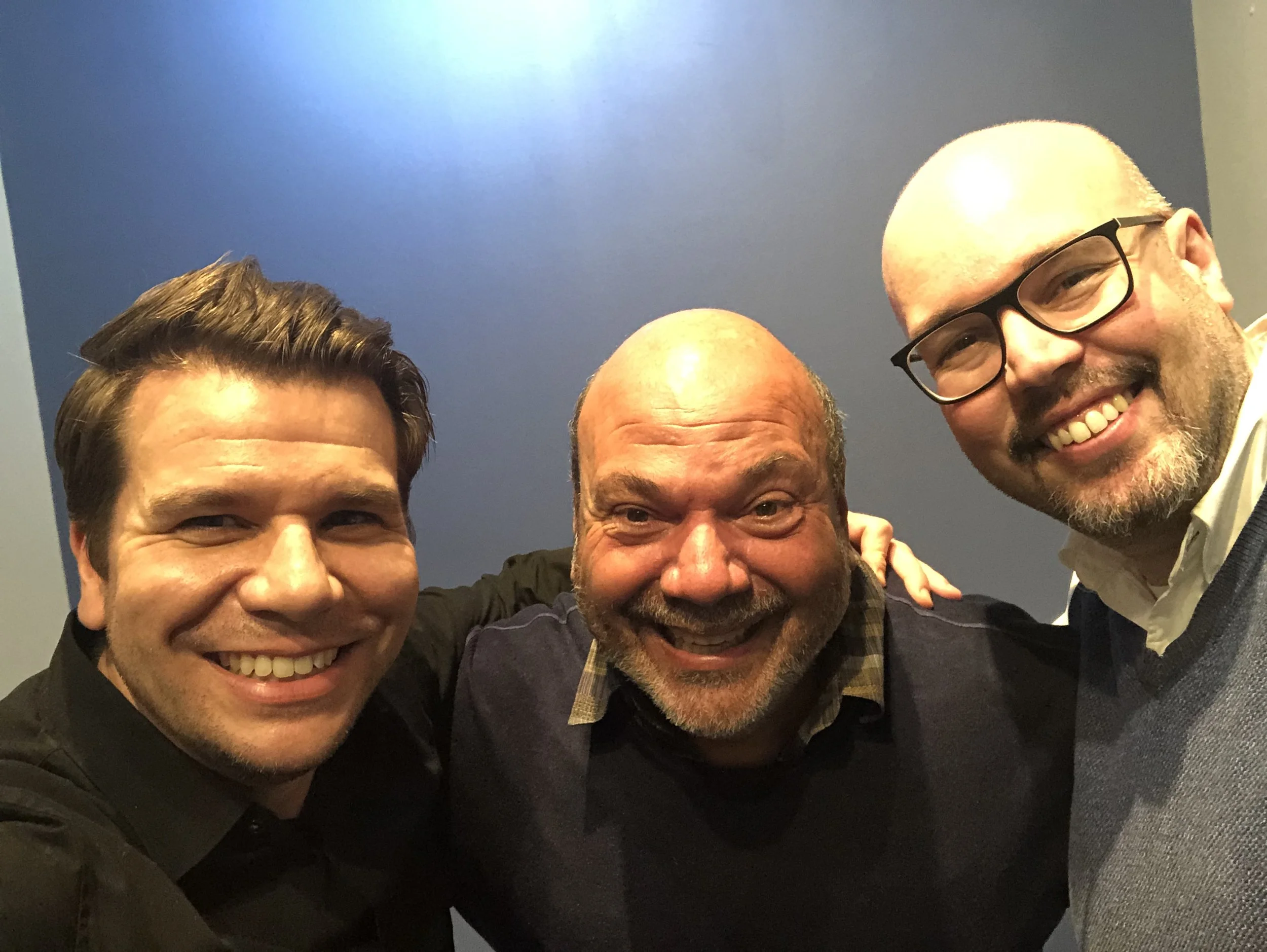 Three smiling men taking a selfie in front of a blue background.