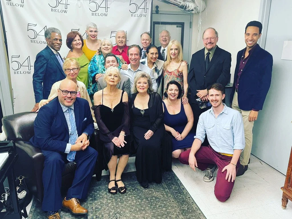Group of 16 people, both men and women, gathered together in an office or studio space for a photo. They are smiling and dressed in business or semi-formal attire. The background has a white backdrop with '54 BELOW' logos.