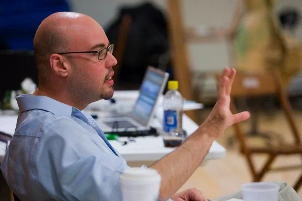 A man with glasses and a bald head sitting at a table with a laptop and a water bottle, gesturing with his right hand in a discussion or presentation.