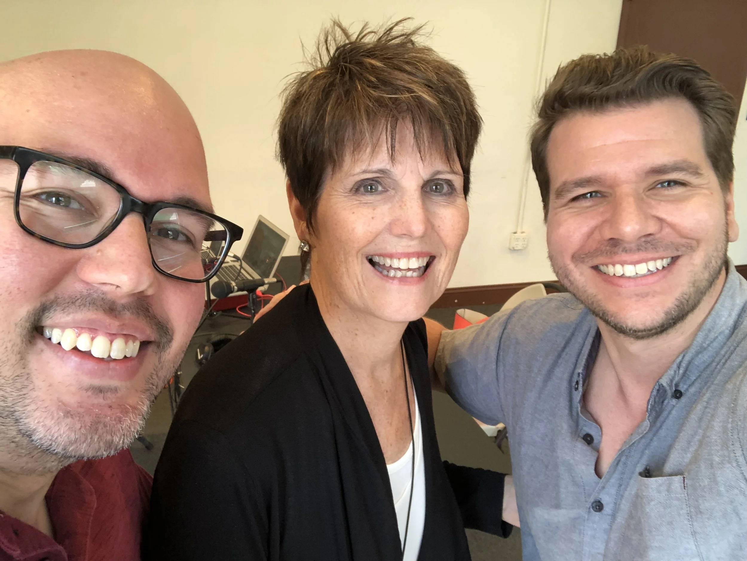 Three smiling people taking a selfie together in an indoor setting with a beige wall and a laptop in the background.