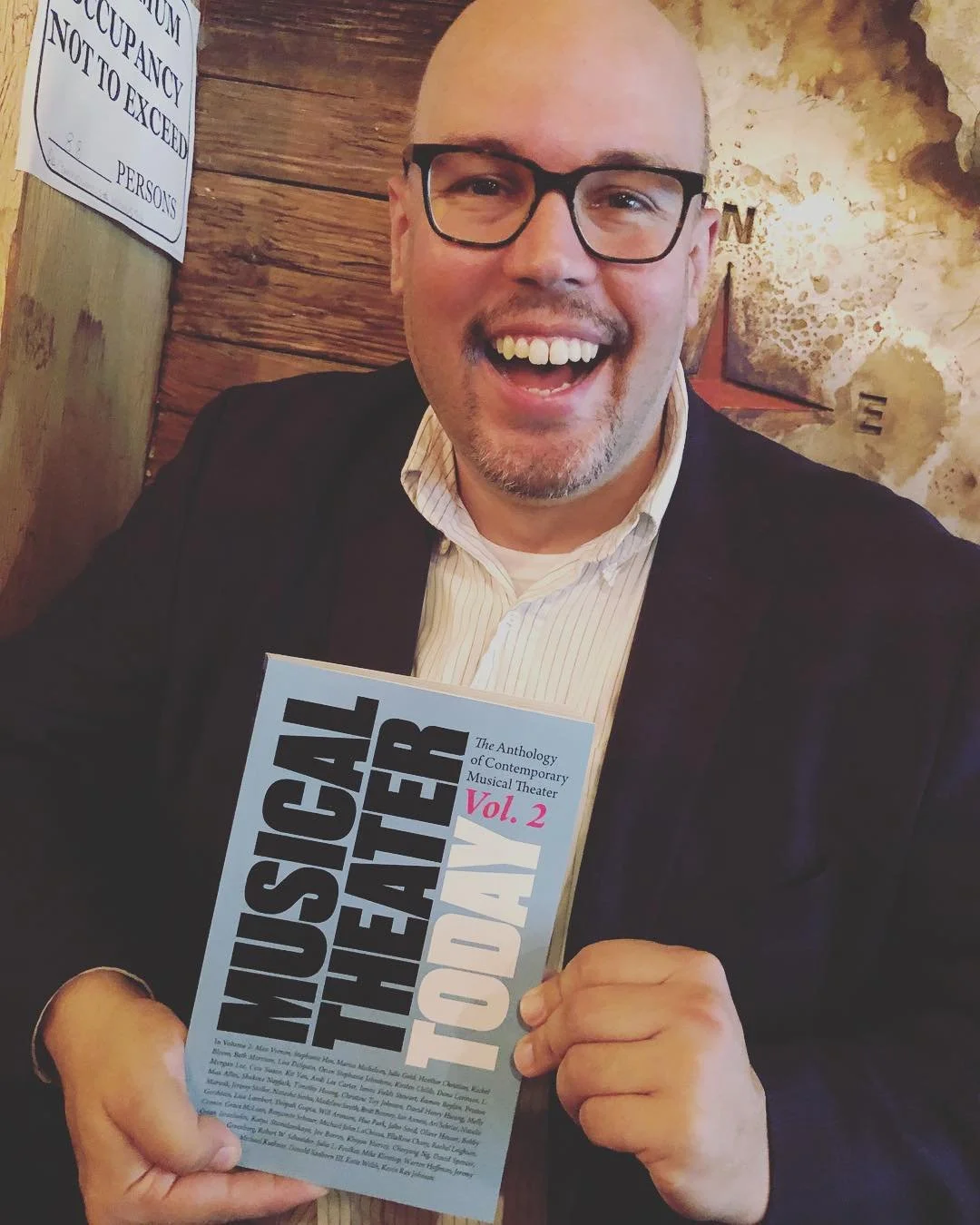 A smiling man with glasses, holding a book titled "Musical Theatre Today Vol. 2" in a cozy, rustic setting with a wooden wall and a decorative arrow pointing north in the background.