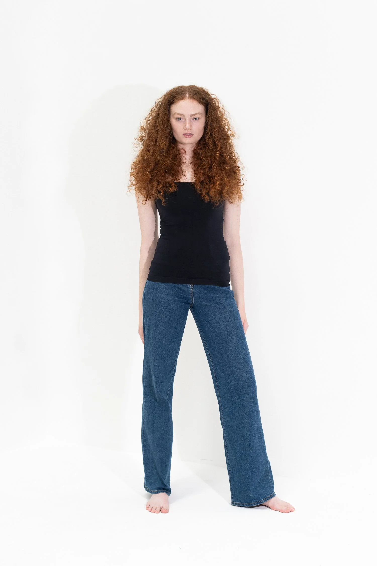 A woman with long curly red hair wearing a black tank top and blue jeans, standing barefoot against a white background.