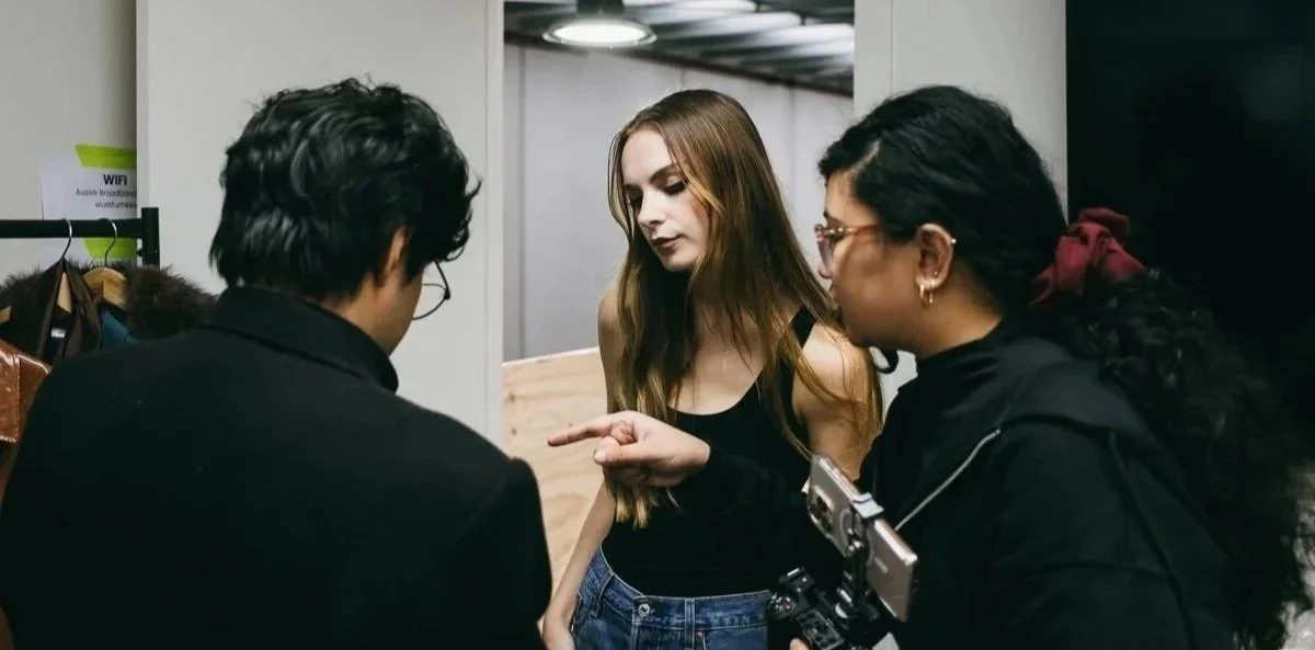 Three women having a conversation indoors, with two women facing away from the camera and the third woman facing side-on, pointing at one of the other women.