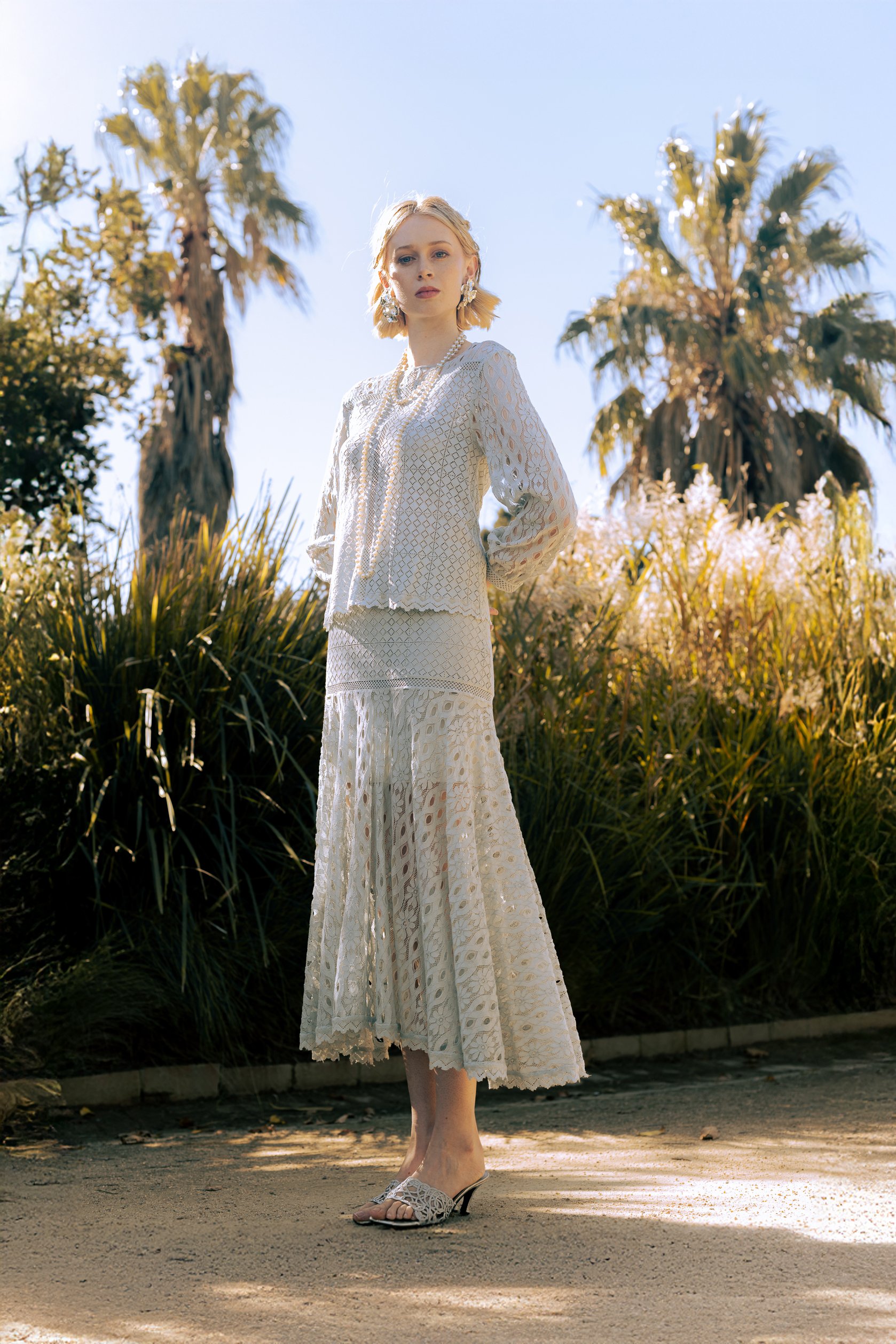 A woman with blonde hair wearing a white lace dress and high heels stands outdoors on a sunny day, with palm trees and blossoming bushes in the background.