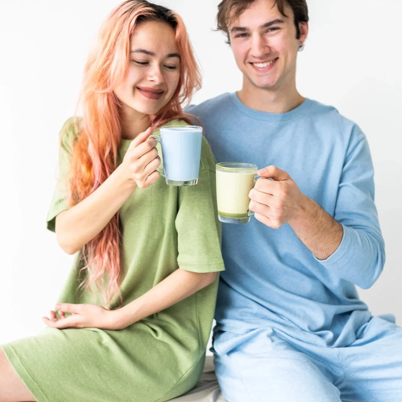 A young woman with pink hair and a young man with brown hair sitting together, smiling, holding glasses with colorful drinks, against a plain white background.