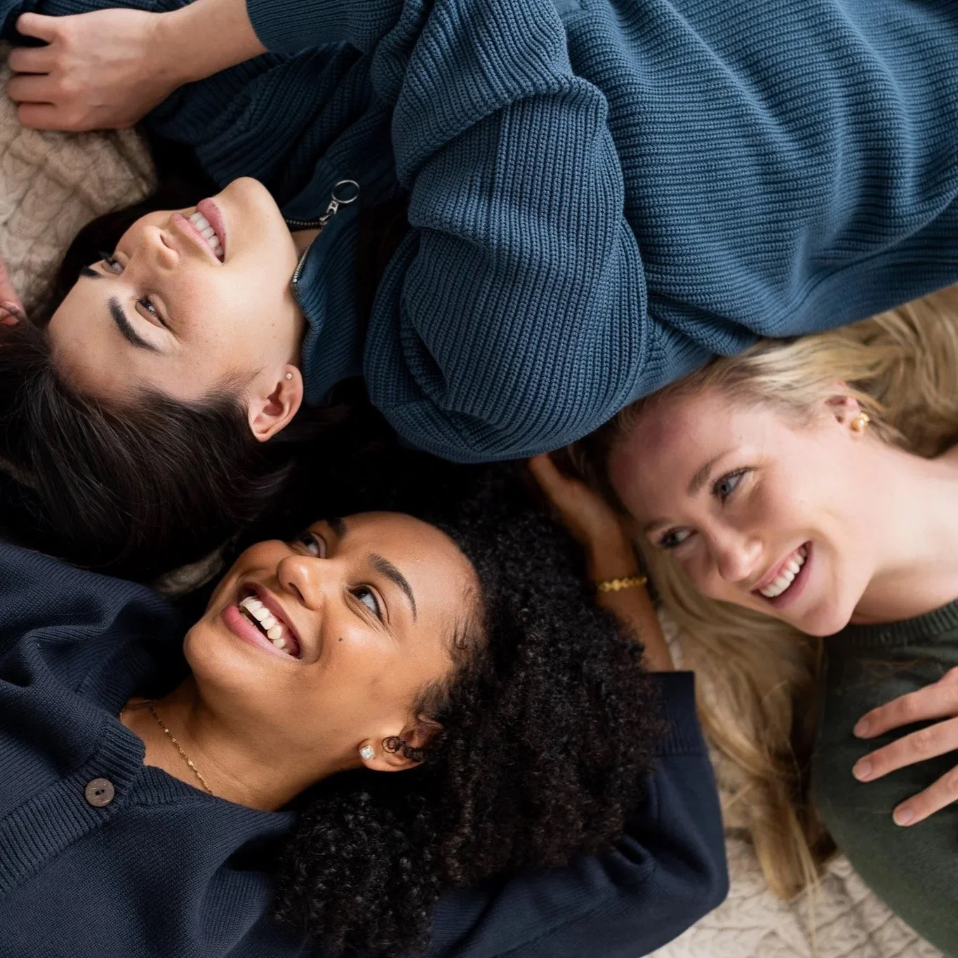 Four women lying on a bed, smiling and looking at each other, in a cozy and relaxed setting.