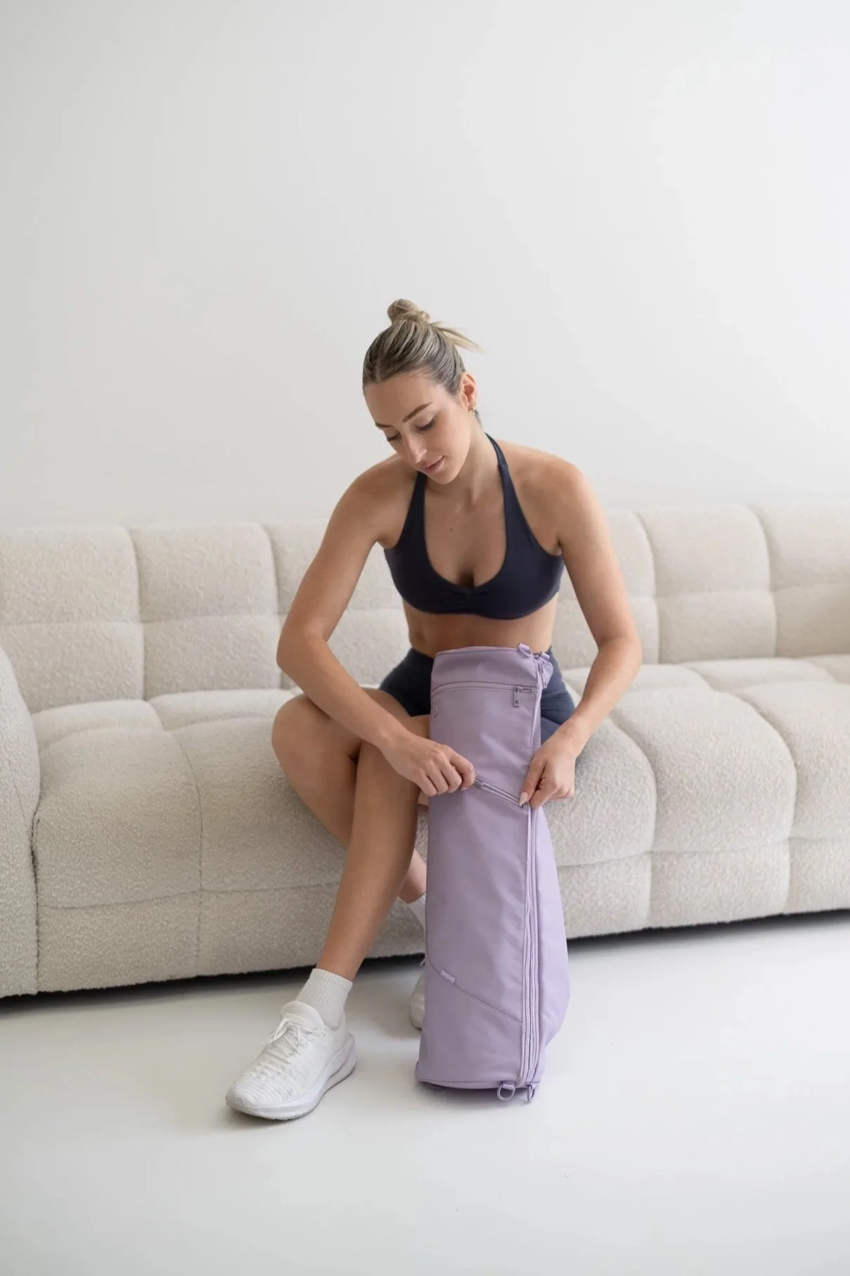 Woman sitting on a beige sofa, putting items into a lavender gym bag, dressed in workout clothes.