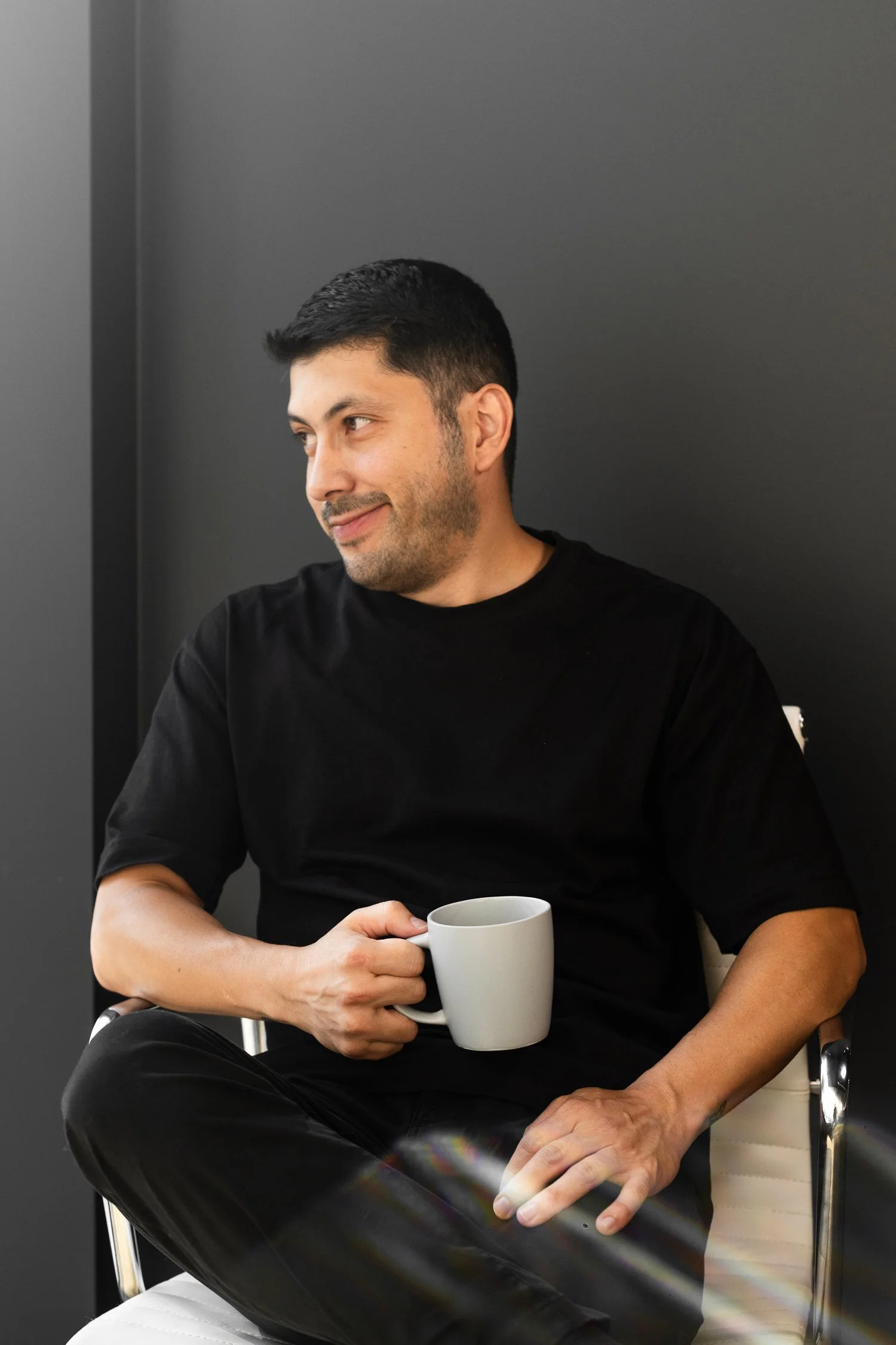 A man with dark hair and a beard, wearing a black T-shirt, sitting against a dark gray wall, holding a white mug, smiling subtly, with his gaze directed to the side.