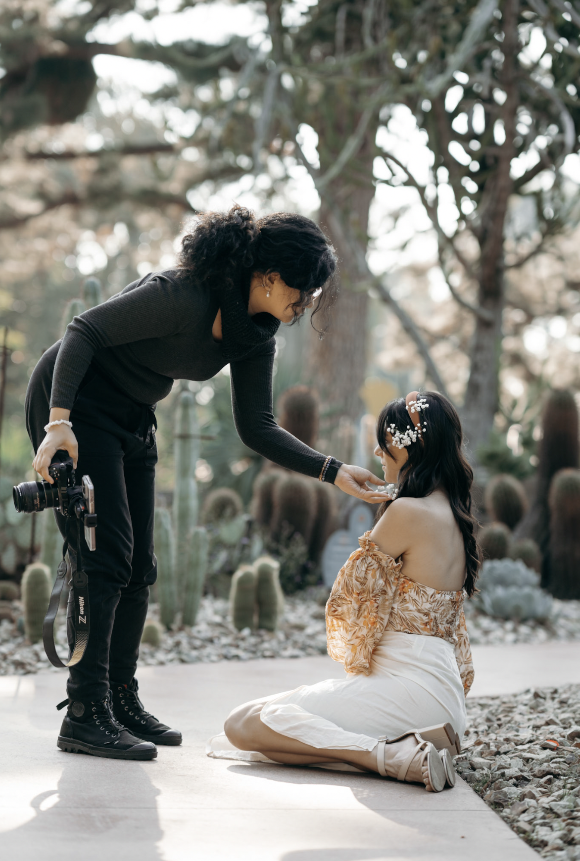 A woman kneels on the ground, wearing a floral off-the-shoulder top and a white skirt, while a photographer leans down to adjust something on her face. The scene is outdoors with plants and trees in the background. Photography session with model.