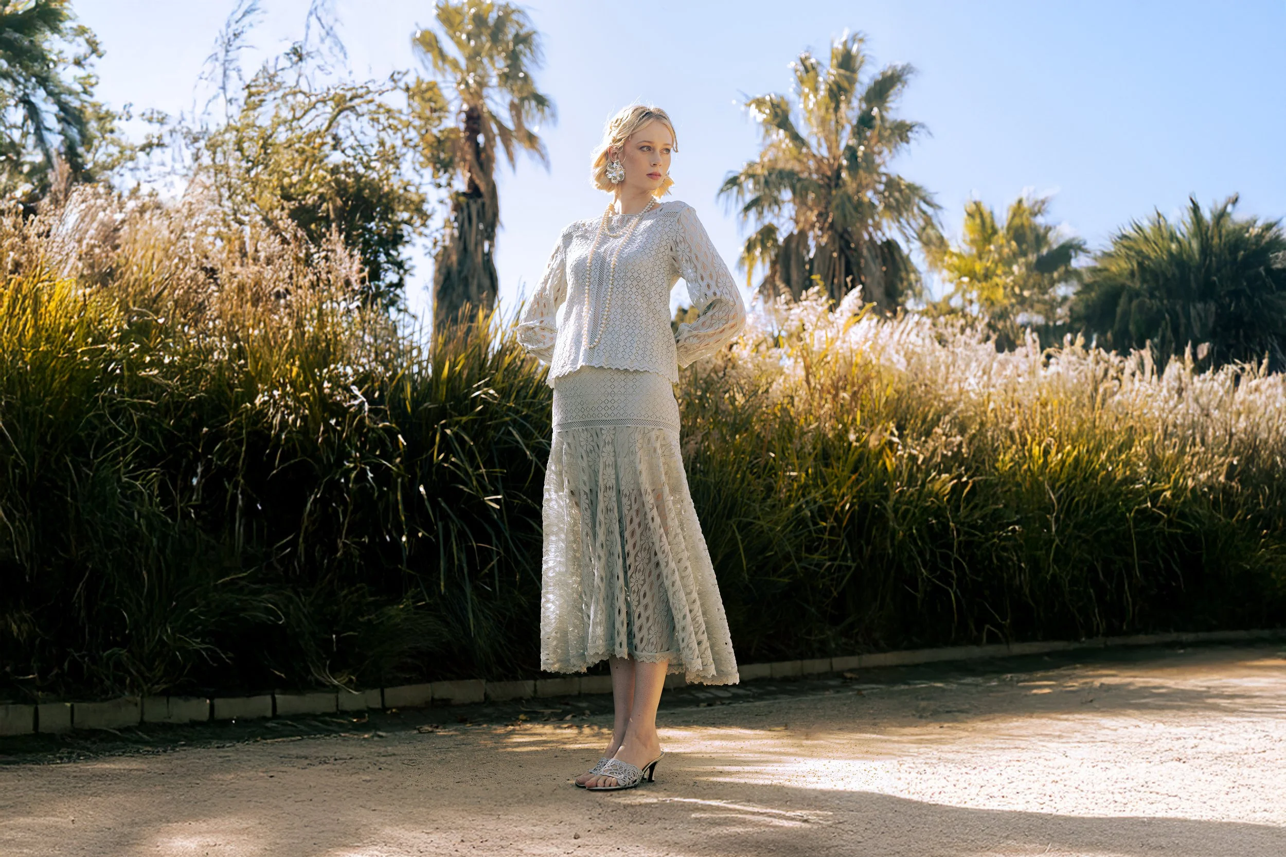 A woman in a white lace dress stands outdoors on a sidewalk, with tall grasses and palm trees behind her. She has blonde hair and is wearing large earrings, a necklace, and high-heeled sandals. The sky is clear and the lighting suggests it is daytime.