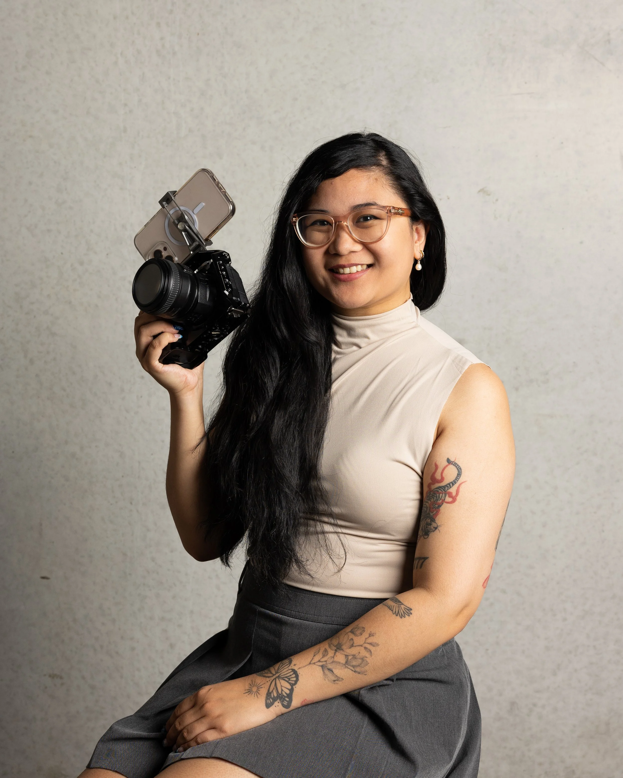 A woman sitting against a plain wall, smiling, holding a camera with a cellphone mounted on it, wearing glasses, a beige top, and gray pants.