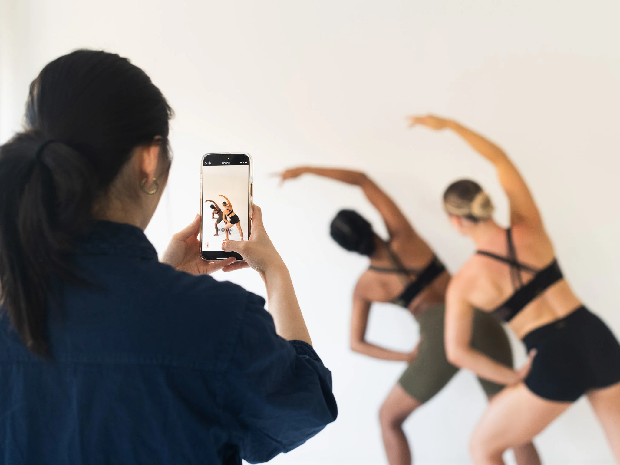 Person with dark hair taking a photo of dancers in black outfits with arms raised on a plain background. Photography session with models. Talent sourcing