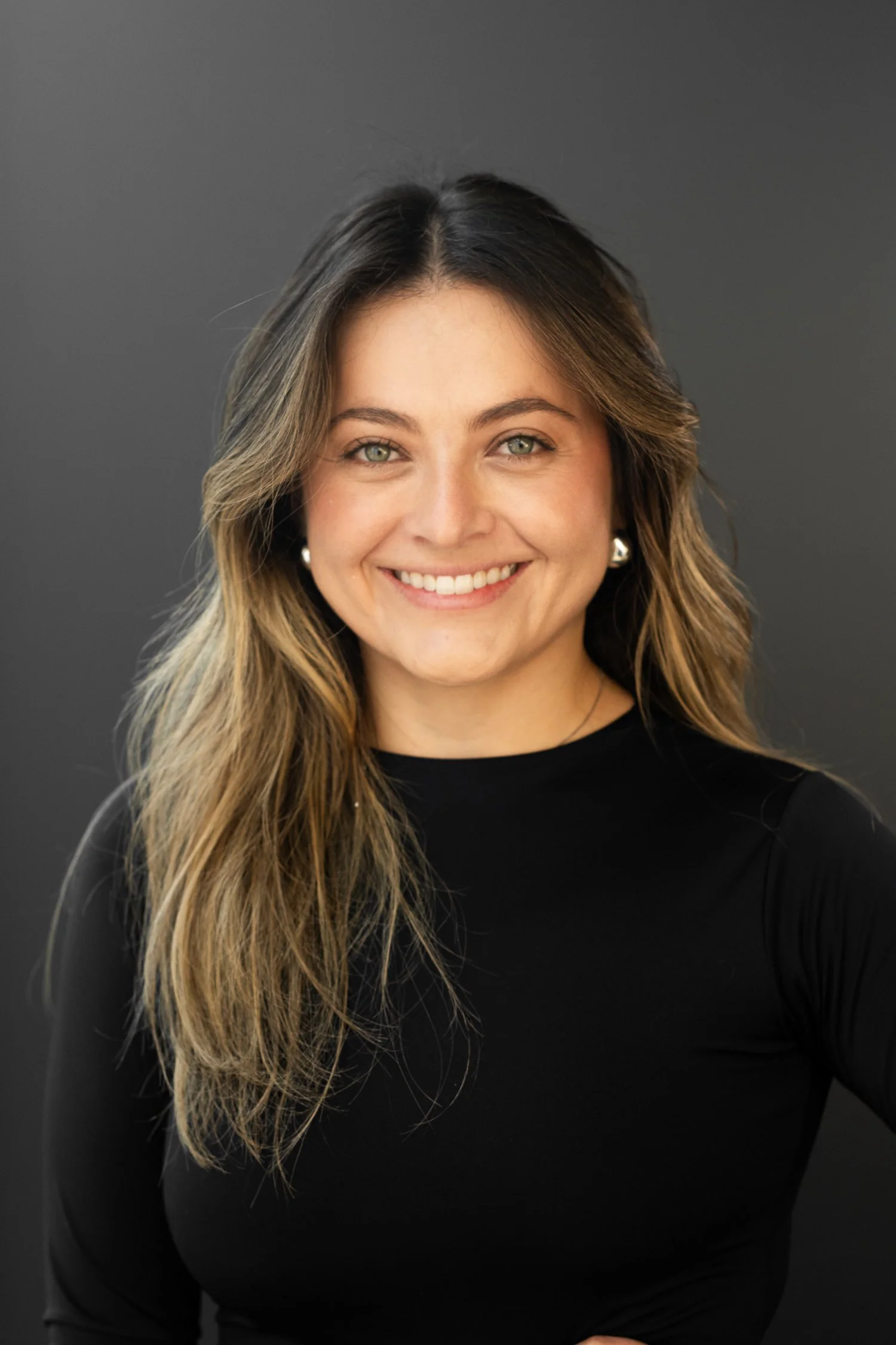 Close-up portrait of a woman with medium-length wavy hair, green eyes, and pearl earrings, smiling against a dark background.