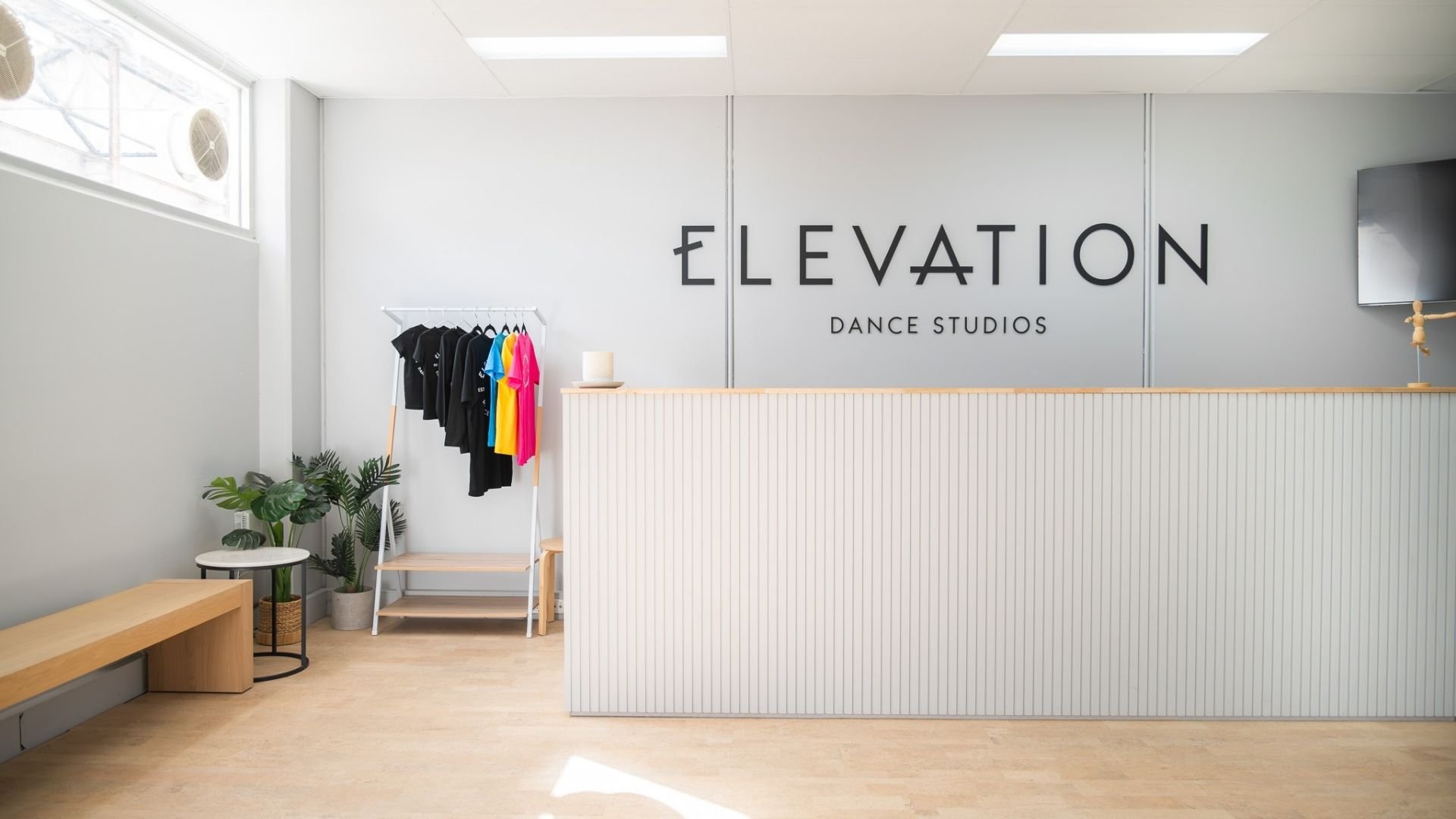 Interior of a dance studio reception area with a large sign that reads 'ELEVATION Dance Studios'. There is a rack of black and colorful T-shirts, a small table with a plant, and a white wainscot panel on the reception counter.